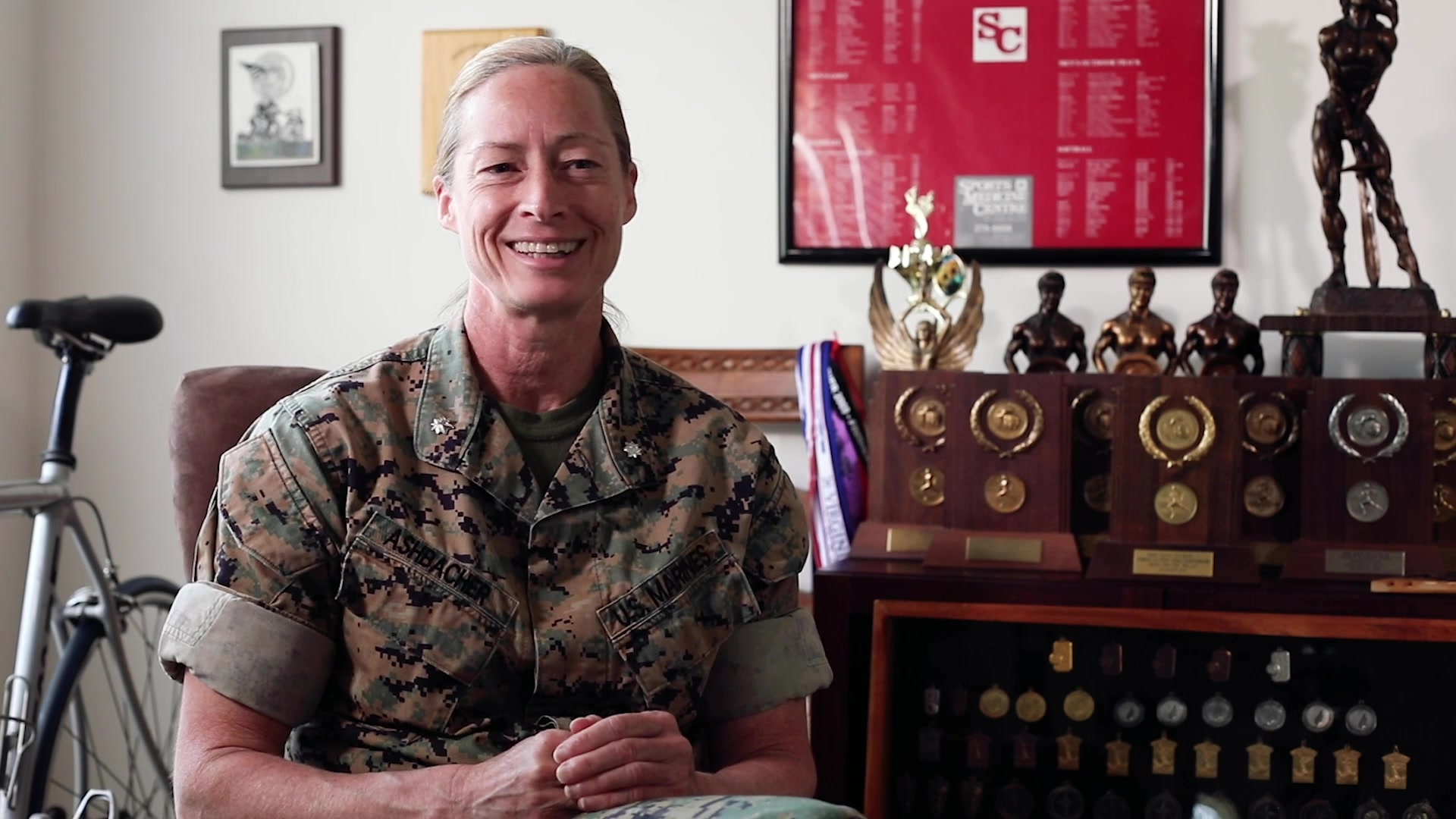 A Marine smiles while sitting and talking in a room in front of trophies and awards.