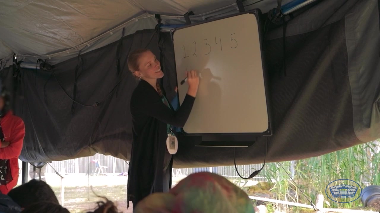 Civilian stands and writes on whiteboard in tent, as seated people watch. 