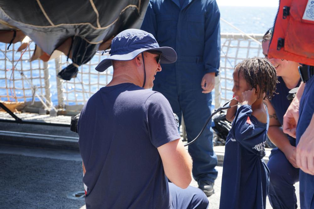 A Coast Guard Cutter Campbell medical crew member lets a child listen to his heart using a stethoscope in the Windward Passage, May 9, 2022. The people were repatriated to Haiti on May 11, 2022. (U.S. Coast Guard photo by Petty Officer 3rd Class Erik Villa-Rodriguez)